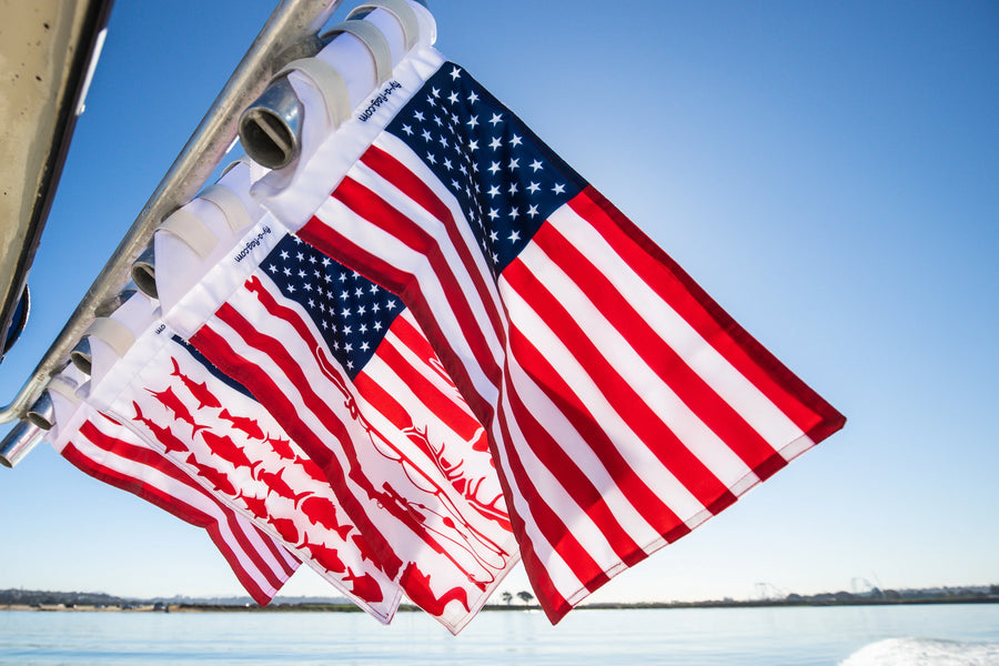 Four flags in a row — American, fishing, and hunting prints — strapped to a boat rail against blue sky