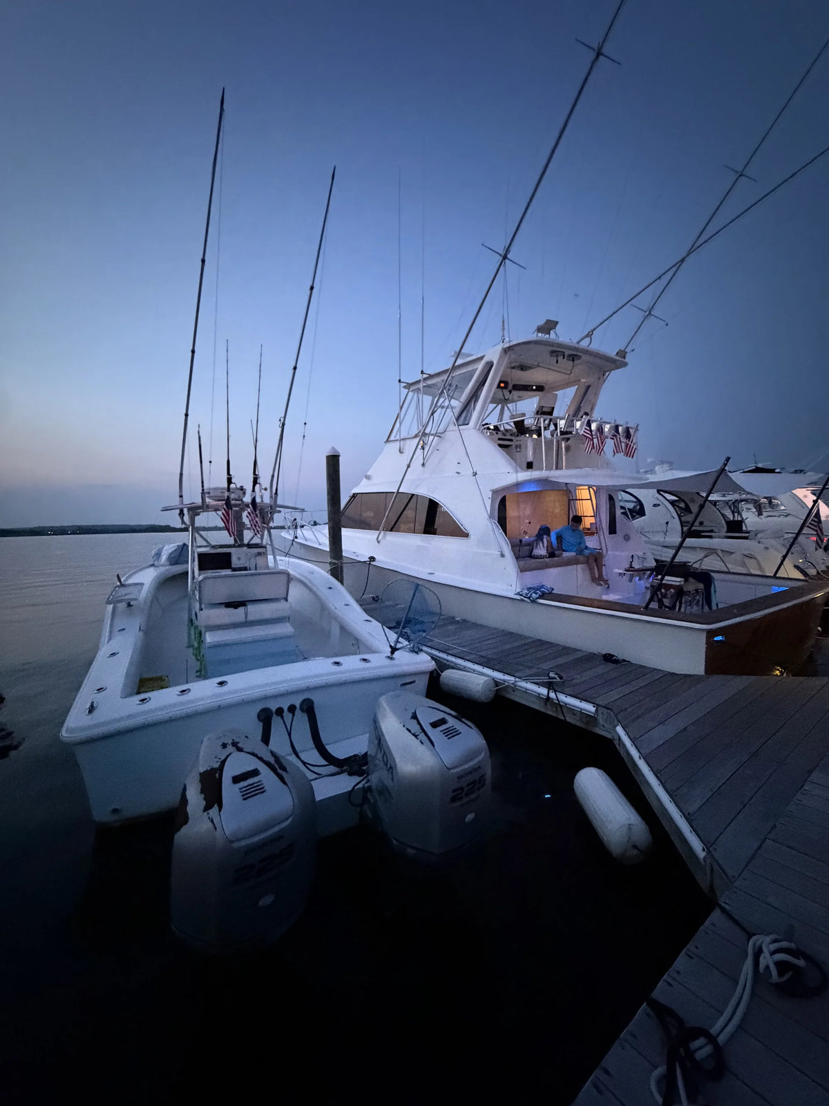 American flags strapped along the bridge rail of a sportfishing boat at dusk, a couple sitting in the lit cockpit