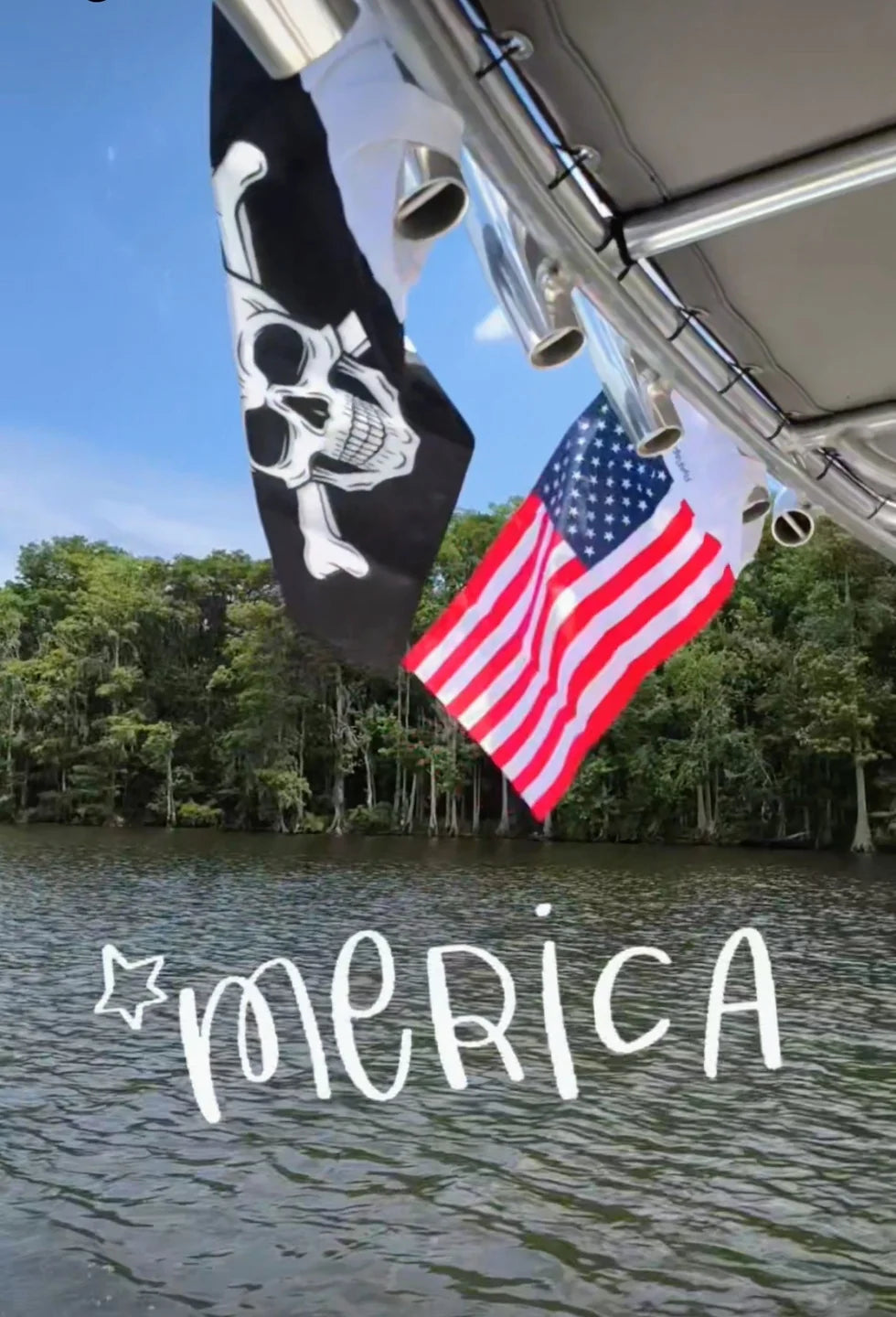 A Jolly Roger and an American flag strapped side by side to a boat T-top over a cypress river, with the word Merica handwritten across the water