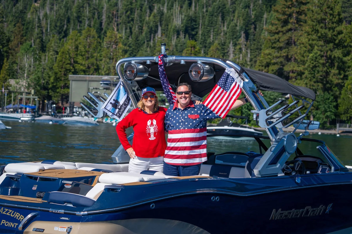 A couple standing in a MasterCraft on an alpine lake, holding an American flag strapped to the tower