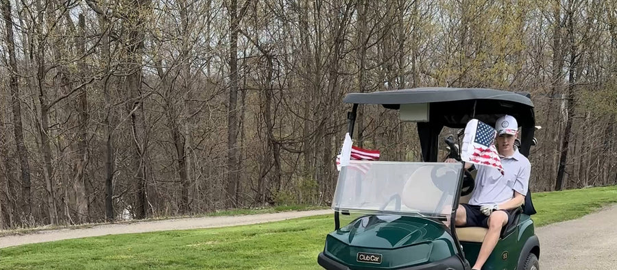 Two American flags strapped to the canopy of a golf cart on a wooded path, rider waving