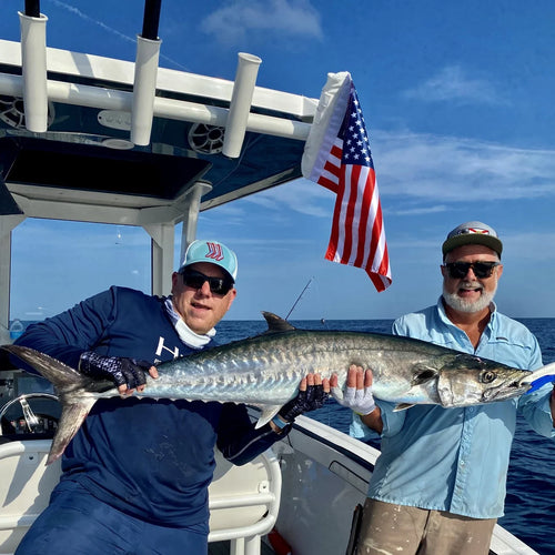 Two anglers holding a king mackerel on a center console, an American flag strapped to the T-top behind them