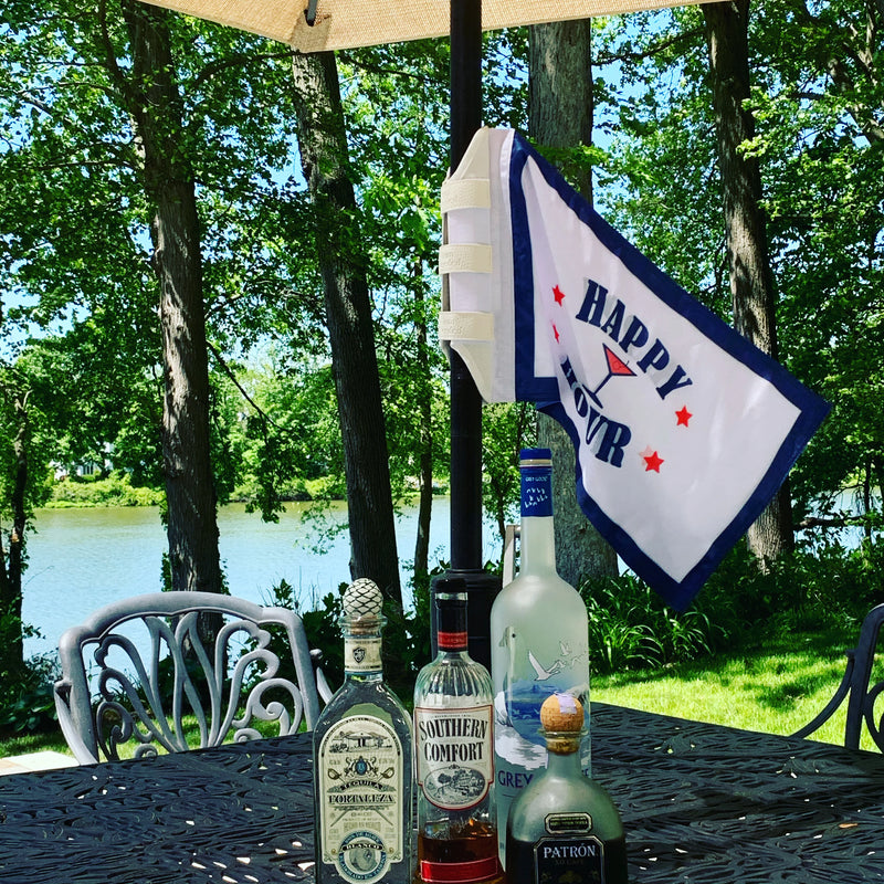 A navy-and-white Happy Hour flag strapped to a patio umbrella pole above a lakeside table