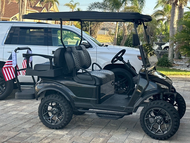 Two American flags strapped to the rear rail of a lifted black golf cart on a paver driveway, palms behind it
